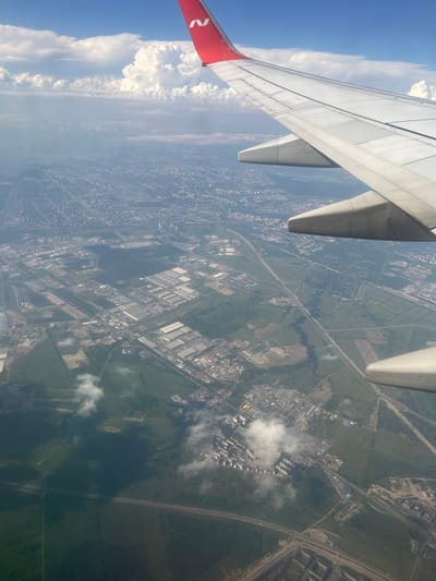 This is an aerial photograph taken from an airplane window, looking down at a landscape that appears to be a mix of urban development and green spaces, likely in Stolbishche, Russia. The view is dominated by the wing of the aircraft in the foreground, painted white with a red section at the tip bearing a white logo. The wing extends diagonally across the image from the upper left to the lower right.

Below the wing, the scene unfolds with a sprawling cityscape characterized by numerous industrial or commercial buildings with light-colored roofs, arranged in a grid-like pattern. These are interspersed with areas of green fields and some bodies of water, possibly rivers or lakes, that snake through the landscape. Roads and what appear to be highways crisscross the area, connecting different parts of the development and the surrounding countryside.

The time of day appears to be daytime, with bright sunlight casting shadows. The weather seems fair, with scattered white clouds visible both in the sky and casting shadows on the ground, suggesting a partly cloudy sky. The overall impression is of a densely populated and developed region undergoing expansion, with a clear demarcation between industrial zones and natural or agricultural land. No people are visible in the image. The visible text is the logo on the airplane wing.