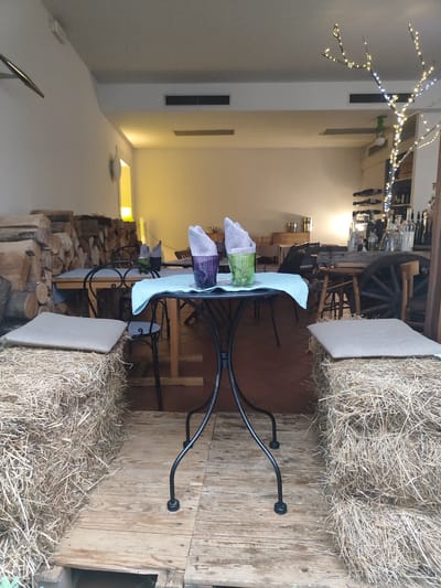 The image depicts an outdoor seating area of a restaurant or cafe, potentially located in Orta San Giulio, Italy, given the rustic and natural elements. In the foreground, two large bales of hay are used as seating, each topped with a light-colored cushion. A low wooden platform serves as the floor for this seating arrangement.

The central focus is a small, round metal table with a black frame. On the table, a light blue cloth is laid out, and on top of it sit two glasses with napkins. One glass is purple, and the other is green, both containing folded grey napkins.

Behind the table, several wooden tables and chairs are visible, some with similar light blue cloths. To the left of the seating area, a large stack of cut logs is piled up, suggesting a rustic or rustic-chic aesthetic.

In the background, the interior of the establishment is dimly lit, with shelves stocked with bottles, likely wine bottles, and a decorative branch adorned with small white and yellow lights. A large wooden cart wheel is also visible. The overall ambiance is casual and inviting, with a blend of natural materials and lighting. There are no people visible in the image.