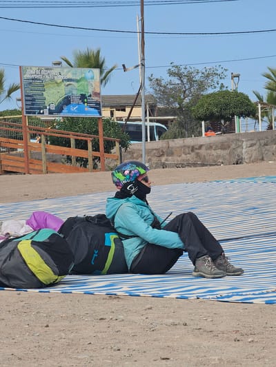 A person wearing a colorful helmet, blue jacket, and hiking boots is seated on a blue and white striped surface, which appears to be laid out on a concrete or stone surface. A large backpack with green, gray, and black elements is positioned behind the person. The background shows vegetation, buildings, and a clear blue sky. A sign with the words "ZONA DE DESPEGUE PARAPENTE ALTO HOSPICIO" is visible, suggesting that the location is a paragliding takeoff zone in Alto Hospicio, Chile. The sign also includes safety regulations and emergency contact information. A fence is present between the sign and the seated person.