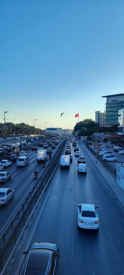 This image depicts a congested highway in Bakırköy, Turkey, during what appears to be late afternoon or early evening. The sky is a clear, vibrant blue, suggesting fair weather. The scene is dominated by a long line of vehicles, indicating a traffic jam. Cars and vans of various colors are visible, mostly in shades of white, black, and silver, filling multiple lanes of the road. A few motorcycles are also present, navigating through the traffic.
In the background, modern buildings stand tall, with one large structure featuring the text "IME İMKB BORSA İSTANBUL" which likely refers to the Istanbul Stock Exchange. Other signage visible includes "METROPORT" and directional signs with Turkish text, confirming the location. A bright red flag is prominently displayed on a flagpole, adding a splash of color to the scene.
The perspective is from an elevated viewpoint, looking down onto the highway. The overall atmosphere suggests a typical urban commuting scenario, with the long queues of cars reflecting the daily grind of city life. The lighting indicates the sun is low in the sky, casting long shadows. There are no visible people clearly identifiable as subjects, as they are mostly inside vehicles. The focus is on the urban landscape and the vehicular traffic.