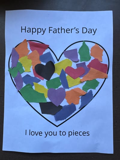The image shows a Father's Day card with a heart shape filled with torn pieces of colored paper. The words "Happy Father's Day" are written at the top of the card, and "I love you to pieces" are written at the bottom. The card is on a dark, textured surface, which is likely a table or floor. There are no people visible in the image. The card appears to be handmade, with the ripped paper pieces creating a mosaic effect within the heart outline. The colors of the paper pieces include orange, yellow, green, purple, blue, red, and black. There are also two smaller heart shapes, one red and one purple, placed within the larger heart. The lighting suggests this photograph was taken indoors, with light coming from above. The location is not discernable from the image.