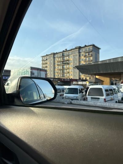 A view from inside a car shows a multi-story building with yellow balconies under a bright sky. A line of white vans is parked alongside the road. The car's side mirror reflects a truck with the word "fresh" visible. Part of the car's dashboard is visible, with sunlight shining on it.