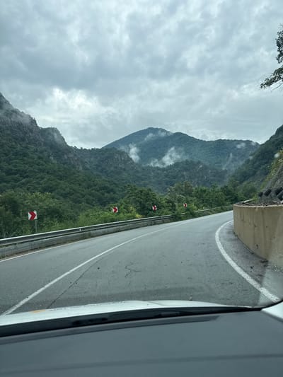 The image shows a curved road winding through a mountainous landscape under a cloudy sky. The road has a white dividing line and a guardrail on the left side. On the right side, there is a concrete wall and some debris. The mountains are covered in dense green trees, and some mist is visible in the valleys. There are two chevron signs on the left side of the road, indicating a curve. The perspective is from inside a vehicle, looking out through the windshield. The scene appears to be a scenic mountain road, possibly in a rural area. No specific details about the city of Krichim, Bulgaria, can be ascertained from the image itself.