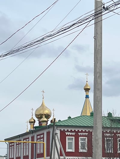 Captured in Норья, Russia, the image presents a partial view of a building exterior, likely a religious structure, characterized by its distinct red and white patterned facade, green roof, and golden domes topped with crosses. The sky is overcast, suggesting a cloudy day. Foregrounded are utility poles and a dense cluster of power lines, obstructing the direct view of the building. The building's architectural details suggest Eastern European influence, contrasting with the urban infrastructure of power lines.