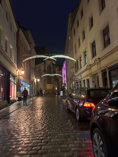 The image captures a narrow, wet cobblestone street in Meissen, Germany, at night or late evening. The street is lined with multi-story historic buildings featuring numerous windows. The cobblestones are highly reflective, indicating recent rain.
Above the street, several decorative white string lights are strung across, forming elegant, arching patterns that resemble stylized birds or waves. In the distance, a prominent building, possibly a church or castle tower, is visible and illuminated with a distinct purple light.
On the right side of the street, a dark-colored car is parked, with its taillights illuminated. Another car is partially visible behind it. Several people are visible on the street, including two more prominent figures in the foreground: one wearing a red jacket and another in a lighter-colored jacket. They appear to be walking along the street. A decorative clock face hangs on one of the buildings on the right.
The scene conveys a quiet, atmospheric evening in a historic town, enhanced by the festive lighting and the glistening wet street. No discernible text is visible on any signs or objects.