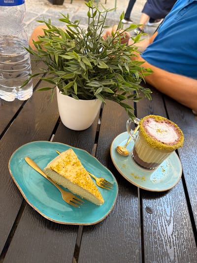 A close-up, overhead shot displays a dark wooden table set with refreshments. A portion of a person wearing a blue shirt is visible on the left, their arm resting on the table. In the center, a clear glass cup filled with a layered beverage, topped with cream and sprinkled with green crumbs, rests on a light blue saucer with a golden spoon. To the right, a slice of light-colored cake, also dusted with green crumbs, sits on a similar light blue plate with a golden fork. A small green potted plant in a white planter is positioned between the drink and the cake. In the foreground on the left, a partially visible plastic water bottle with a blue label is resting on the table. The scene appears to be outdoors, with ample natural light suggesting daytime. The overall ambiance is relaxed and inviting, suggesting a moment of enjoyment at a cafe or restaurant, possibly in Valletta, Malta, given the potential context. The sunlight creates subtle reflections on the polished table surface, highlighting its textured grain. The focus is on the food and drink, with the background slightly blurred.