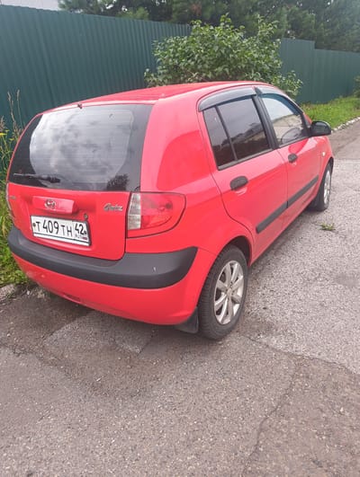 A red Hyundai Getz is parked on a gravel and asphalt surface next to a green metal fence. The car is viewed from the rear left side. The license plate, clearly visible, reads "T 409 TH 42" with "RUS" below it. The car has tinted windows and a rear wiper. Green foliage and trees are visible behind the fence, suggesting a semi-urban or rural environment. The ground surface is rough with visible gravel and some cracks in the asphalt. The lighting suggests it is daytime, with some cloud cover reflected in the car's rear window. No people are present in the image.
