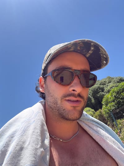 A tanned man with a light beard and mustache, wearing sunglasses and a patterned cap, looks upward towards a clear blue sky. A white towel is draped over his shoulder, and a silver chain necklace is visible. Green foliage is partially visible in the background. The man's posture and the angle of the shot suggest he may be lying down.