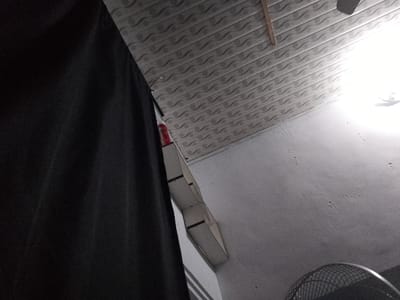 This is a low-angle, wide shot of an indoor space with a textured ceiling and a white wall. A large, dark fabric object, possibly a curtain or clothing, occupies the left side of the frame, obscuring much of the scene. In the upper right quadrant, a bright light source illuminates the ceiling and wall, with a portion of a ceiling fan visible. Attached to the wall, a narrow, box-like structure with a segmented appearance is visible. There are no people or animals present in the image. The scene does not provide any specific indicators of its location within Senti, Nigeria.