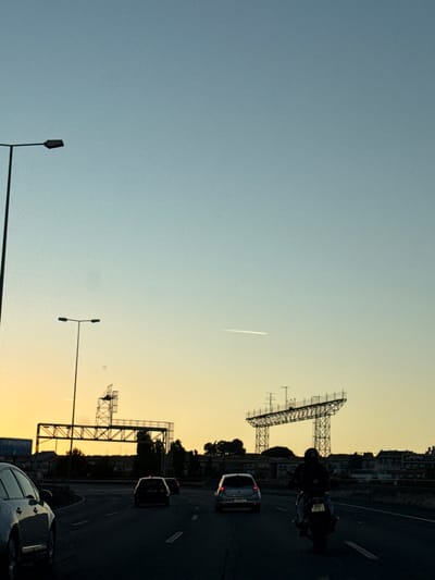 This image captures a scene on a highway in Loures, Portugal, during what appears to be sunset. The sky transitions from a pale blue at the top to a warm yellow and orange hue near the horizon. A distant airplane contrail is visible in the sky. On the left side of the road, a tall street lamp stands prominently.
In the middle ground, a large, industrial-looking metal structure, possibly a billboard support or part of a construction project, dominates the view. Below it, trees and buildings form a silhouette against the colorful sky.
The foreground shows a highway with several vehicles. A silver car is on the left, partially visible. Several other cars are driving on the road, including a dark-colored car in the lane to the right of the silver car, and another car further ahead. On the right side of the highway, a motorcyclist, wearing a helmet, is riding a motorcycle. The road surface is dark asphalt, with visible lane markings. The overall impression is a busy highway scene as day transitions into evening.