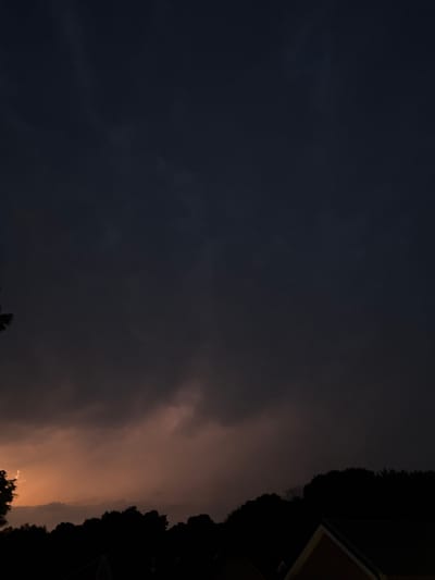 The image shows a stormy sky with dark clouds and a hint of orange light from the horizon. Silhouetted against the sky is a line of trees and the rooftops of buildings. The location is Putten, Netherlands. There are no people visible in the image. The scene is dark and overcast, suggesting it is either dawn, dusk, or during a storm.