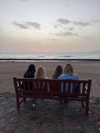 The image depicts four individuals seated on a maroon bench facing a beach and the ocean at sunset. The sky is a gradient of pale yellow and pink, with scattered clouds. The ocean is calm with gentle waves. The beach is sandy with some rocks. In the background, there are a few other people visible on the beach and some buildings on the horizon. The ground in front of the bench is paved with dark stones. This scene is likely located on the coast of Arona, Spain, given the general appearance of the beach and ocean.