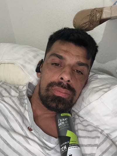 The image features a close-up, angled portrait of a man lying down, likely on a bed or couch, in an indoor setting in Oberasbach, Germany. He has dark hair, a beard, and a mustache, with his eyes open and a neutral to slightly somber expression. He is wearing a light-colored t-shirt with grey stripes. A black earpiece is visible in his left ear.
A prominent object in the foreground is a can of "Dove Men+Care Advanced Invisible Fresh" deodorant, resting near his head. The visible text on the can includes "TRIPLE ACTION TECHNOLOGY," "INVISIBLE FRESH," "12H PROTECTION," "ANTI WHITE MARKS," and "ANTI YELLOW STAINS."
In the upper left corner, an object resembling a light brown shoe or sandal, partially covered in brown tape, is visible. The man is resting on a white pillow with subtle grey stripes, and white bedding is also visible around him. The activity depicted is simply the man relaxing or resting. The lighting suggests an indoor environment, but the specific time of day is not discernible.
