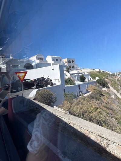 This media file captures a view of a hillside village with whitewashed buildings and narrow streets, characteristic of Santorini, Greece. The scene is bathed in bright sunlight, suggesting a clear day. The architecture features rounded arches and cubic structures, with some buildings perched precariously on the steep incline. Vegetation, mostly dry and scrubby, dots the landscape.

In the foreground, the reflection in what appears to be a car window shows a portion of the road and a yellow triangular warning sign with an image of falling rocks or debris. Behind the sign, several motorcycles are parked. A dark-colored car is also visible, partially obscured by the reflection. The overall impression is one of a sunny, Mediterranean day in a picturesque, possibly tourist-oriented location. The orientation of the image is sideways.