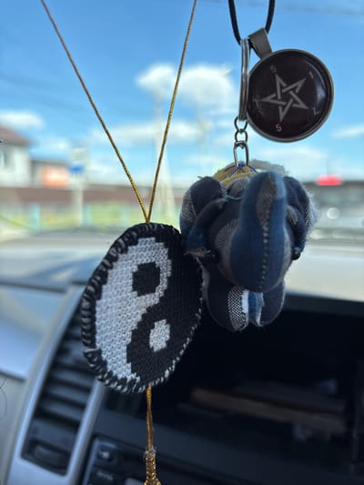 In Chigiri, Russia, three small ornaments hang from a car's interior, visible through the windshield. The leftmost ornament is a yin and yang symbol, intricately knitted in black and white. To its right, a small stuffed elephant made of striped blue and gray fabric is attached, with black beads for eyes. Above these, a circular pendant hangs, featuring a white five-pointed star or pentagram on a dark background, with numbers "5," "1," "2," and "5" arranged around it. The ornaments are suspended by thin golden and black cords against a backdrop of a bright blue sky with scattered clouds and a blurred view of buildings and traffic, suggesting an outdoor scene observed from within a vehicle.