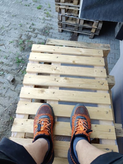 The image shows a pair of orange and black athletic shoes standing on a wooden pallet structure. The shoes are laced with orange laces and appear to be new or well-maintained. The pallet structure is made of unfinished wooden planks, some of which are reinforced with plastic sheeting underneath. The ground around the pallets is a mix of dirt, gravel, and scattered patches of grass. In the background, several other wooden pallets are stacked neatly. To the right of the pallets, a dark gray, textured surface is visible, possibly a wall or a large object. The lighting suggests it is daytime, with shadows indicating an outdoor setting. The overall scene is utilitarian and appears to be in an area where materials are stored or processed. The prompt's request to include the location of Łomna-Las, Poland, cannot be fulfilled as there are no visual cues in the image to indicate this specific geographical location.