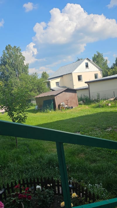 The image depicts a residential property in Nemėžis, Lithuania, on a sunny day with scattered white clouds in a blue sky. In the foreground, a green railing is visible, partially obscuring a garden bed with red and yellow roses and other white flowers, bordered by a dark wooden fence. Beyond the garden is a well-maintained lawn.
Further back on the property, a weathered, brown metal shed with a corrugated roof stands in the middle of the yard. Next to the shed, a green metal fence panel is propped up, and a wheelbarrow is present. A brown, two-story house with a light-colored facade and a silver metal roof is situated behind the shed. This house has multiple windows, including a dormer window on the upper level. To the right of this house, another light-colored building with a sloped roof is visible. Trees, including a birch tree, and dense green foliage surround the buildings and yard. The overall scene suggests a rural or suburban residential setting.