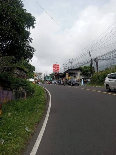 The scene depicts a two-lane asphalt road curving slightly to the right under an overcast sky in Saing Ta Mawt Lay, Myanmar. The road's left edge is marked by a solid white line, adjacent to a grassy verge containing scattered plastic litter and a concrete retaining wall with purple graffiti. Further left, a large tree with dense green foliage obscures a portion of the hillside, with utility lines and a street light pole visible.
On the right side of the road, a silver/white SUV (Mitsubishi Xpander model) is partially visible at the far right, moving away from the viewer. Further down the road on the right, a line of buildings, likely commercial establishments, features corrugated metal elements and signs. A prominent red billboard for "PRINGSEWU" is visible, displaying text such as "Pringsewu Family," "Lele Bakar," and "KUFTE." Below it, a green sign with "TALITA" is partially seen.
Multiple utility poles with a dense network of black overhead wires line the right side of the street. Traffic congestion is visible further down the road, with several motorcycles stopped or parked near the buildings, and a black sedan alongside them. Additional vehicles, including a silver/grey SUV, are present in the distance. Several indistinct individuals are visible near the parked motorcycles on the right. In the background on the right, dark green, heavily vegetated hills rise under the cloudy sky. A large truck is visible on the far left of the road in the distance, facing the viewer.