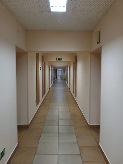 The image shows a long, straight, and well-lit interior hallway. The hallway has beige walls and a beige, textured ceiling with a long rectangular fluorescent light fixture. The floor is tiled with large, square, light-colored tiles forming a path down the center, bordered by a narrower strip of brown tiles along the sides. On the left side of the hallway, there are several doorways and what appear to be vertical wooden panels on the wall. A green emergency exit sign with a running figure is visible on the wall at the far end of the hallway, as well as on the left wall closer to the foreground. There is also a green exit sign at the very end of the hallway on the right. At the far end of the hallway, there appears to be a wider opening or a junction, with a blue sign visible. The perspective creates a strong sense of depth, drawing the viewer's eye towards the end of the corridor. The environment appears to be a public building or facility. It is not possible to determine the specific location in the city of Progress, Russia, from the image alone.