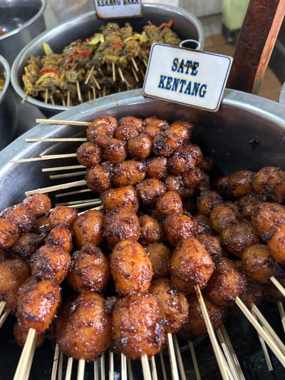 The image shows a close-up view of several skewers of "Sate Kentang" (potato satay) in a large metal container. The potatoes are small, round, and appear to be glazed with a dark, sticky sauce, some pieces showing charring from grilling. They are arranged on wooden skewers. A white sign with blue text reading "SATE KENTANG" is visible in the upper right portion of the image. Behind the potato satay, a similar metal container holds skewers of what appears to be "Kerang Dara" (blood clam) satay, identifiable by the darker, less uniform shapes and specks of red and yellow ingredients. The scene is likely a food stall or market in Cirebon, Indonesia, a city known for its diverse street food offerings, particularly satay variations. The lighting suggests an indoor or covered outdoor setting, possibly a market during daylight hours. There are no people visible in the frame.