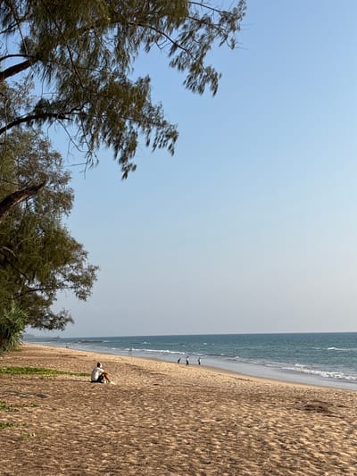 The image captures a beach scene in Khok Kloi, Thailand. The sky is clear and light blue. The beach, composed of textured sand, occupies the right side of the frame. A person wearing a white shirt and dark pants is visible on the beach. The ocean is located along the upper edge of the sand. To the left of the frame are trees.