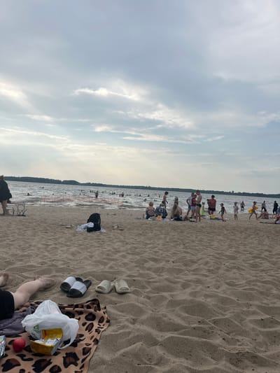 A wide shot captures a crowded beach on a cloudy day. The foreground features a sandy expanse with several pairs of sandals, a leopard-print blanket, a plastic bag with snacks, and an apple. Further back, the water is filled with people swimming and wading, with a temporary barrier demarcating a swimming area. On the shore, groups of people are gathered, some sitting on towels or chairs, while others stand and converse. The background shows a treeline across the water, indicating a natural landscape. The lighting suggests overcast conditions, with diffused sunlight breaking through the clouds in places. The overall impression is a lively, communal summer day at a lakeside or seaside resort.
