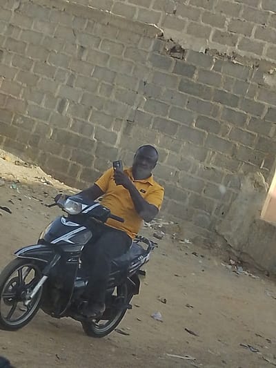 A man in a yellow shirt sits on a black motorbike in front of a cinder block wall. He holds a mobile phone up, possibly taking a photo or recording a video. Debris litters the ground next to the wall. The man faces the camera with a smile. The area appears to be a dirt or sand lot.