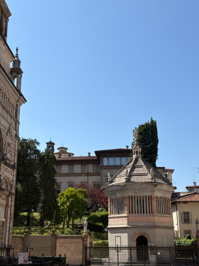 The image captures a daytime scene in Mozzo, Italy, likely in an urban or historical district. In the foreground on the left, a portion of a richly decorated building facade, characterized by intricate carvings and arches, is visible. To the right, a distinct octagonal structure, possibly a baptistery or mausoleum, stands prominently. This structure features a colonnaded upper section and is topped with statues and a spire. Behind these, several trees, including a tall cypress, are interspersed with buildings displaying terracotta-tiled roofs and classical architectural elements, such as windows with shutters. A clear blue sky dominates the upper portion of the image. A low stone wall with inscriptions and a metal fence are present in the lower foreground. There are no discernible people or actions in the visible portion of the image.