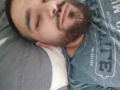 A close-up, low-angle selfie shows a young man lying on his side, his head resting on a pillow. The man has dark hair, a beard, and is wearing a gray t-shirt with white lettering. The background consists of a pillow with a gray and white striped pattern. The image appears to be taken indoors, possibly in a bedroom. The user has requested to include the location in the city of Vrachesh, Bulgaria, which is not determinable from the provided image.