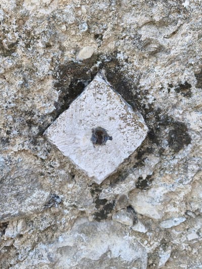 A close-up, overhead shot shows a diamond-shaped, light-colored stone embedded in a rough, greyish rock surface. The stone is smooth and worn, with visible chisel marks. A dark, irregular hole is present in the center of the diamond. The surrounding rock is textured and appears weathered, with patches of dark moss or lichen. The lighting suggests an outdoor environment. This object is a geodetic marker, a type of surveying landmark. This particular marker is located on the Via del Pozzo in the town of Campochiaro, Italy.