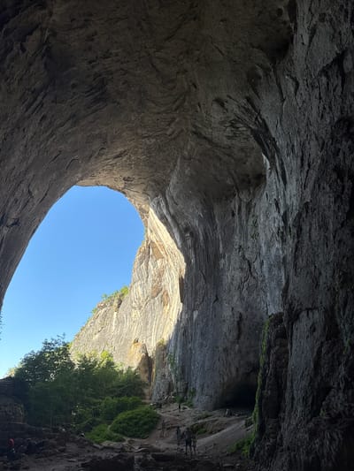 This image depicts the interior of the Prohodna Cave, also known as the "Eyes of God," located near the village of Karlukovo, Bulgaria. The cave is characterized by its massive scale, with towering rock formations forming a natural archway that frames a bright blue sky. Sunlight streams through this opening, illuminating the textured rock faces and casting shadows within the cavern.
The foreground of the image is dark and rocky, with uneven terrain suggesting a natural, unpaved surface. Various people are scattered throughout the scene, some appearing to be exploring or resting. In the mid-ground, a group of individuals is visible near the entrance of another section of the cave, with lush green foliage and trees growing against the cliff face. The rock walls themselves display a rough, layered texture, indicative of geological erosion over time. One rock face in the background shows a prominent, lighter-colored band, possibly a different mineral composition. The overall impression is one of a vast natural wonder, with human presence adding a sense of scale and exploration.