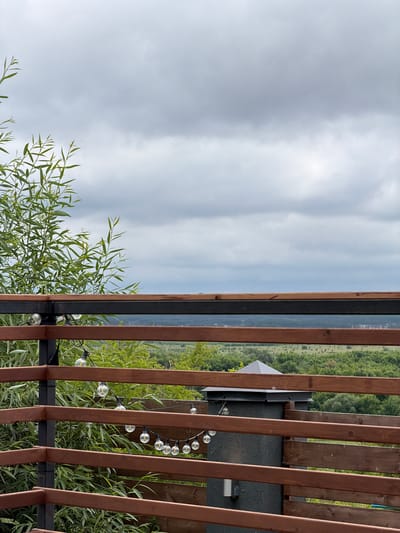 The image depicts a view from a wooden deck, looking out over a verdant, hilly landscape under a cloudy sky. The foreground is dominated by the railing of the deck, constructed with horizontal wooden planks. String lights with exposed bulbs are draped along the railing. To the left, a tall, leafy green plant with long, slender leaves extends upwards, partially obscuring the view. The middle ground reveals a panoramic vista of rolling hills covered in dense green trees, with scattered signs of human habitation, possibly a village or suburban area, visible in the distance. The sky is overcast with varying shades of gray clouds. The scene is set in Семилуки, Russia. There are no people or animals visible in the image.