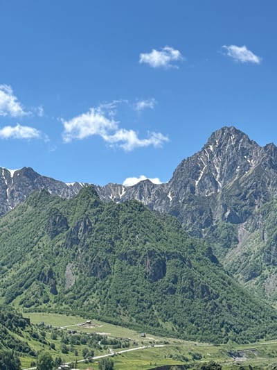 This is a landscape photograph featuring a steep, forested mountainside under a bright blue sky with scattered white clouds. The camera perspective is looking up at the mountain, which is densely covered in green trees. The rugged terrain of the mountain is visible, with rocky outcrops and patches of snow near the summit.

In the foreground, a portion of the sky is visible, with a few wispy clouds. The transition from the sky to the mountain is sharp, creating a dramatic contrast between the blue expanse and the textured green of the trees. Further down the mountainside, a cleared area of green grass can be seen, with a few buildings and a winding road or path visible within it. This suggests a human presence or settlement nestled within the mountainous landscape.

The lighting suggests it is daytime, likely mid-morning or afternoon, given the brightness and the shadows cast on the mountain. The weather appears clear and sunny, ideal for outdoor activities or viewing the scenery. The overall impression is one of a remote, natural environment with a small human settlement integrated into it. The image evokes a sense of awe and the grandeur of nature. There is no visible text in the image.