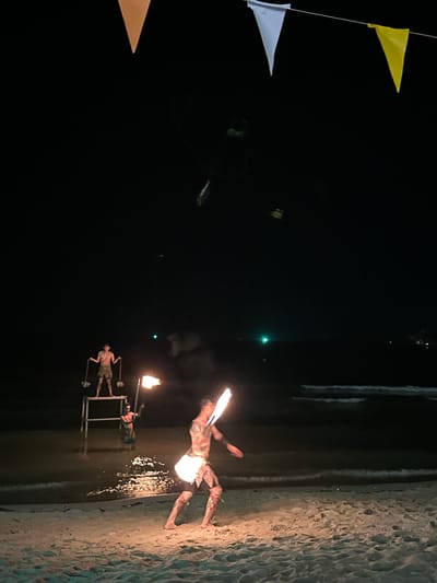 Night scene on a beach in Baan Klong Prao, Thailand. Three triangular flags—yellow, white, and orange—are strung along a line in the upper left corner. Two people are performing with fire on the beach. One person is near the water's edge with two flaming torches mounted on a stand. The other person, covered in tattoos, is lying on the sand with two torches and one in their mouth. Light from the flames reflects off of the wet sand near the water.
