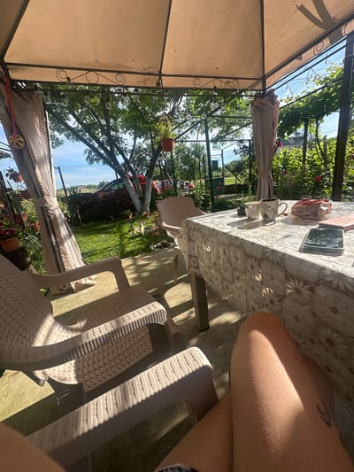 The image depicts a covered outdoor seating area, likely in Rogozen, Bulgaria, during daylight hours. The area features a table draped with a floral-patterned tablecloth, set with two coffee mugs, one of which reads "COFFEE," and a package of biscuits. A smartphone and a small notebook are also present on the table. Two light-colored lounge chairs are positioned in the foreground, facing away from the camera. The background reveals a lush green garden with trees, flowering plants, and a fence. Sunlight streams through the canopy, casting shadows on the ground and furniture. The overall scene suggests a relaxed outdoor setting, possibly a backyard or patio, in a rural or suburban environment.