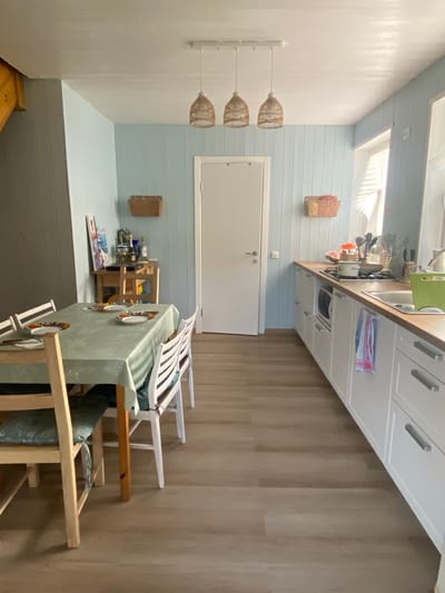 The image shows a bright and airy kitchen and dining area. The walls are paneled with light blue boards, and the floor is covered with light brown laminate flooring. A dining table covered with a light green tablecloth is set with plates and cutlery. Four chairs surround the table, two with light wood frames and cushions, and two with white frames. A small wooden shelf unit with various items is placed near the corner. In the background, a white door is visible. The kitchen counter is lined with white cabinets and a wooden countertop. A stove with a pot is on the counter, along with utensils and other kitchen items. A window with blinds is visible on the right side of the kitchen. Three woven pendant lights hang from the ceiling above the dining area. The scene appears to be a residential kitchen in Leipyasuo, Russia.