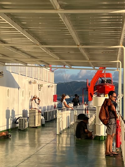 The image captures a scene on the deck of a ferry, likely near Batas Desa Skotong Timur, Indonesia. The deck is made of reflective green material, and a white canopy supported by beams covers the area. Several individuals are present. In the foreground, a man with glasses and a brown backpack stands near a railing, looking towards the right. To his left, a person is sitting on the floor, engrossed in their phone. Further down the deck, a woman in a white top stands by a railing, facing away from the camera. A man in a black jacket is also visible in the background. On the left side of the image, a person in a light-colored outfit is holding a camera, with several air conditioning units mounted on the white wall of the ferry. A red lifeboat davit is prominently displayed in the middle ground. The background features a body of water with distant mountains under a cloudy sky. The lighting suggests it is either early morning or late afternoon, with long shadows cast on the deck.