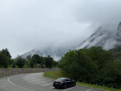 The image depicts a parking area on a cloudy day, likely in the vicinity of Dzivgis, Russia, given the mountainous terrain and vegetation. A black sedan is parked in one of the marked spots on the asphalt. The parking lot is curved, bordered by a stone wall and lush green grass. Behind the wall, trees and a gentle slope lead up towards a dense forest. In the background, misty mountains rise, partially obscured by clouds. The overall atmosphere is serene and natural.