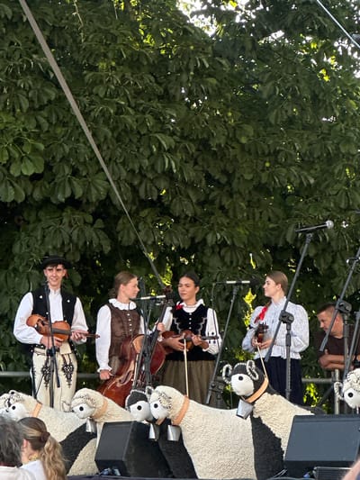 The image shows a group of young people performing music on an outdoor stage, set against a backdrop of lush green trees. Four individuals, two males and two females, are prominently featured in the center of the frame, dressed in traditional attire. The male on the left wears a black hat, a white shirt with black trim, and a dark vest. He is holding a violin and appears to be playing it. Next to him, a female wears a white blouse with a ruffled collar and a brown vest. She is positioned behind a cello, holding its bow. Another female, in the center, wears a black vest with white trim over a white shirt and is holding a ukulele. To her right, a female is dressed in a light-colored blouse with floral patterns and a dark skirt, holding a violin. In the foreground, several large, plush sheep figures with bells around their necks are placed on the stage, facing the audience. To the far right, a man with short hair is visible, leaning against a railing. Microphones and speaker equipment are set up on the stage. The overall atmosphere suggests a cultural event or performance taking place in a park or outdoor venue. The presence of traditional clothing and musical instruments indicates a focus on folk heritage. The backdrop of trees suggests the performance is taking place outdoors in a natural setting. The image is taken from a slightly low angle, looking up at the performers.