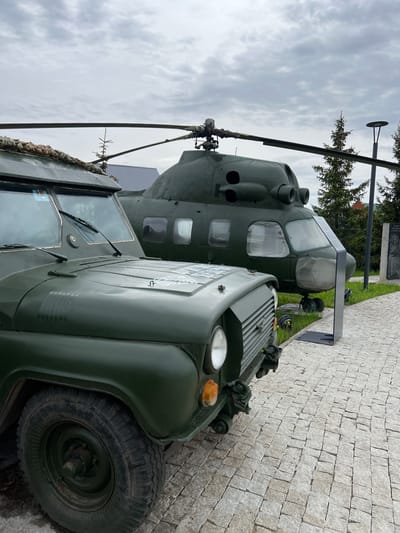 The image shows a Soviet Mil Mi-24 helicopter and a UAZ-469 military vehicle, both painted in olive drab, exhibited outdoors in the city of Введенская Слобода, Russia. The helicopter features its characteristic tandem cockpit, twin exhaust nozzles, and a prominent rotor blade. The UAZ-469, with its utilitarian design, includes a spare tire mounted on the hood and a canvas covering. The vehicles are displayed on a paved area with a cloudy sky in the background.