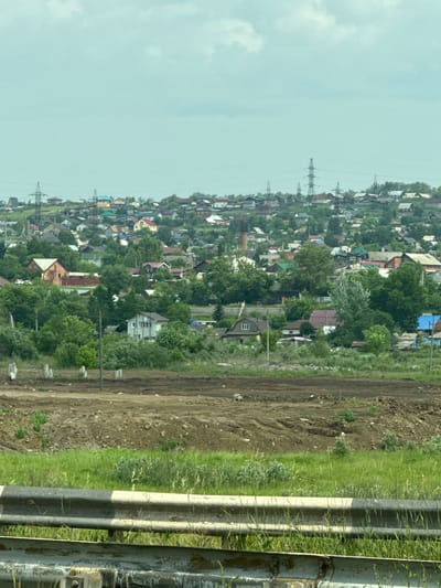 The image depicts a suburban hillside community in Solontsy, Russia, with a clear sky and scattered clouds overhead. A dense collection of houses, varying in size and architectural style, covers the undulating terrain. Many of the houses are single-family dwellings with pitched roofs, some painted in vibrant colors like red and blue, while others are more muted. Green trees and foliage are interspersed throughout the residential area, suggesting a well-established, vegetated neighborhood. Prominent in the middle ground are several tall, lattice-like electricity pylons, indicating the presence of power infrastructure. In the foreground, a section of a highway barrier, painted with alternating black and white bands, separates the viewer from a grassy embankment and a cleared, dirt construction site with visible tire tracks and piles of soil. The overall scene suggests a developing or expanding residential area on the outskirts of a city. There are no people visible in the image.
