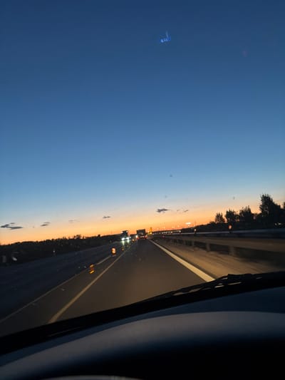 The image depicts a view from inside a vehicle driving on a highway at dusk. The sky is a deep blue, transitioning to orange and yellow near the horizon, indicating the sun has recently set. There are a few scattered clouds. In the distance, there are lights from other vehicles, streetlights, and what appears to be a power line or bridge structure. The road has lane markings, and a highway barrier is visible on the right side. The windshield of the vehicle is in the foreground, with the wipers partially visible. There is also a distant object in the sky that resembles an aircraft. The scene is characteristic of a highway in or near Bicske, Hungary, during twilight hours.