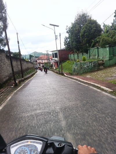 The image depicts a street scene in Masaiti, Zambia, from a first-person perspective, looking forward over the handlebars of a moving motorcycle or scooter. The road, appearing wet or damp, is asphalt and slopes downhill into the distance, showing motion blur in the foreground.
Several motorcycles, each with a single rider, are visible ahead, traveling down the street in the same direction.
To the left, a tall, grey concrete or stucco wall runs along the roadside, partially obscured by utility poles carrying multiple overhead wires. Vegetation is visible behind the wall further down the road.
To the right, a reddish-brown sidewalk parallels the road. Beyond it, a prominent green-painted concrete retaining wall with block-like sections is present, featuring white graffiti, including the letters "AFP," near its base. Above this wall, a grassy slope ascends to dense green trees and foliage. A tall metal utility pole with a solar-powered street light stands on the right. Further down the street on the right, buildings with light-colored walls and reddish-orange tiled roofs are visible, one appearing to be a commercial establishment with signage.
In the background, under an overcast sky, vegetated hills or mountains rise, with additional houses featuring red-tiled roofs nestled among them. The immediate foreground shows the upper portion of a motorcycle's dashboard, including a speedometer and a driver's right hand gripping the handlebar.