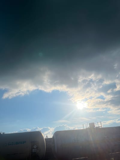 This is a vertical, medium-angle shot of a cloudy sky with the sun shining brightly through the clouds. The sky is a mix of bright blue and darker, overcast areas, suggesting a change in weather. Wispy white clouds are illuminated by the sun, creating a dramatic effect. On the right side of the image, two train tank cars are partially visible, their metallic surfaces reflecting the light. The train cars appear to be stationary.

The time of day is likely afternoon, given the bright sun and its position in the sky. The weather is dynamic, with patches of clear sky and heavy cloud cover. The presence of train cars suggests an industrial or railway setting. The overall mood is somewhat dramatic and awe-inspiring due to the interplay of light and shadow in the sky.

Visible text on the train cars includes what appears to be Cyrillic lettering. On one car, the numbers "57872830" and "20" are clearly legible. Another car has text that includes "ЗАО «СИБУР-ТРАНС»". There are also hazard symbols and other markings. The location context provided is Kauchuk Stantsiya, Russia.