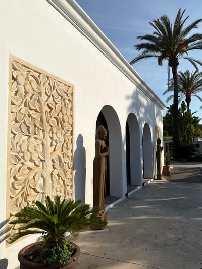 The image captures a sunlit exterior of a white building with arched entrances, likely a structure in Sant Joan de Labritja, Spain. A stone relief depicting a fruit tree adorns the wall to the left. Two stone statues of figures with hands clasped in prayer stand in front of the building, one closer and more prominent, the other further back. A potted palm plant is in the foreground. Palm trees are visible in the background, with a clear blue sky above. The ground is paved with concrete, and shadows cast by the building and palm trees create a geometric pattern.