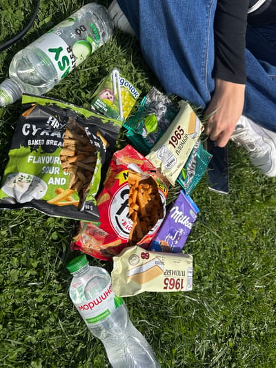 The image shows a person sitting on grass in Sîngera, Moldova, surrounded by snacks and drinks. The person is wearing jeans and white sneakers, and holding a smartphone. The assortment of food and beverages includes: two open bags of "HRUS TEAM" Baked Bread Snacks, one with "SOUR AND" flavor, and the other "BAC" Wheat-Rye Crisps; sunflower seeds; juice box (ДОЧОК) with apple and grape flavor; two "1965 ПЛОМБІР" ice cream bars, and a Milka chocolate bar. There are also two bottles of mineral water, labeled "EASY WATER" and "Моршинська". The background consists of a green grassy area with small clover.
