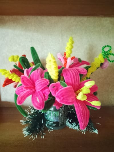 The image shows a close-up, eye-level view of a craft arrangement of colorful pipe cleaner flowers. The flowers are predominantly pink with yellow accents, interspersed with green pipe cleaner leaves. They are arranged in what appears to be a small, dark-colored container, possibly a candle jar as indicated by the visible label on the right side of the container which reads "lavender". The container is adorned with a fringe of black and white tinsel or craft material. The arrangement is set against a textured, light-colored wall and rests on a wooden surface, likely a shelf or table. The lighting is soft, casting subtle shadows. The image was taken indoors. There are no people visible in the image. The background and environment suggest a domestic setting, not a specific location in Rheda-Wiedenbrück, Germany.