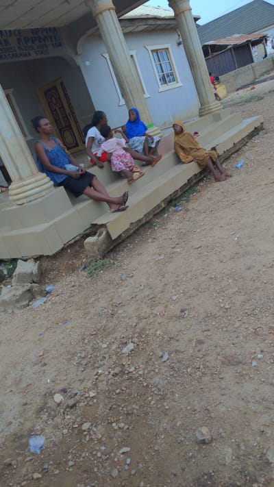 Five people are sitting on the steps of a building. The building has columns and a window with panes. There is dirt and debris on the ground in front of the building. The location is Masaka, Nigeria.