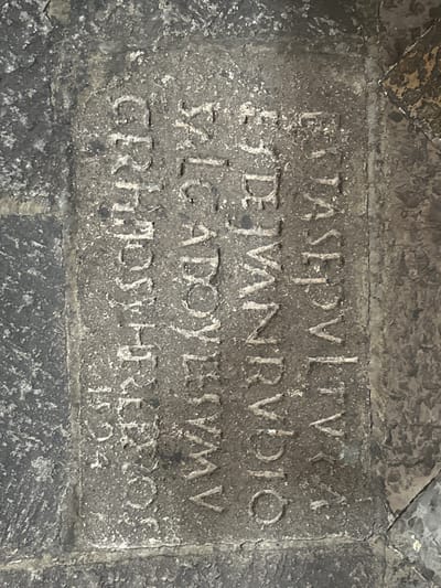 The image displays a weathered stone inscription, likely a historical marker or tombstone, embedded in a textured surface. The inscription consists of several lines of Roman numerals and letters, heavily eroded and difficult to decipher fully. The stone is dark grey and appears to be part of a larger structure or paving. The lighting suggests an indoor or shaded environment, possibly a crypt or an ancient ruin. The overall condition of the inscription indicates significant age and exposure to the elements. The context of the city of Huejotzingo, Mexico, suggests this inscription could be related to the region's colonial history or indigenous heritage, though the specific content is not clearly legible.