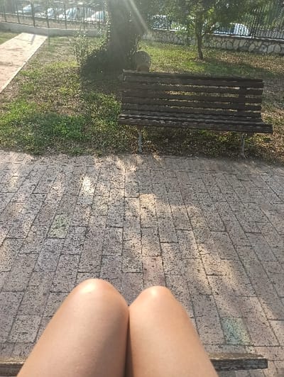 The scene depicts an outdoor public space within San Marzano sul Sarno, Italy, viewed from a first-person perspective of an individual seated on a bench. The precise location within the city is not identifiable. In the foreground, the bare, light-toned upper thighs and knees of the seated individual are visible. Immediately beyond, a textured paved surface, composed of rectangular or square blocks, extends forward under direct sunlight from the upper right, which also causes lens flare. A strip of sparse grass separates the paving from a weathered wooden park bench with metal supports. Behind this bench, a tree trunk and foliage are present. The background features a low stone wall topped with a black metal fence or railing, bordering an area with parked vehicles. The scene is brightly illuminated, indicating daytime conditions. No other individuals are depicted.